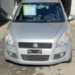 Silberner Suzuki Kleinwagen vor Werkstatt im Sonnenlicht, Ansicht von vorne.