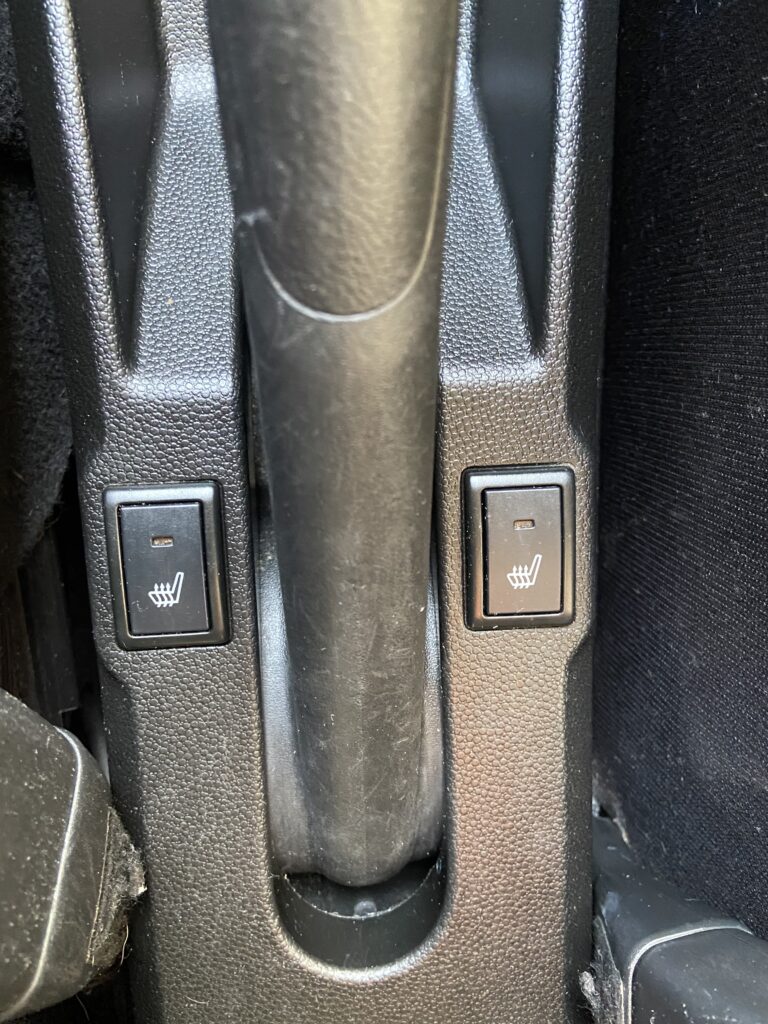 Schalter für Sitzheizung in einem Auto. Links und rechts neben dem Handbremshebel im Fahrzeug-Innenraum.