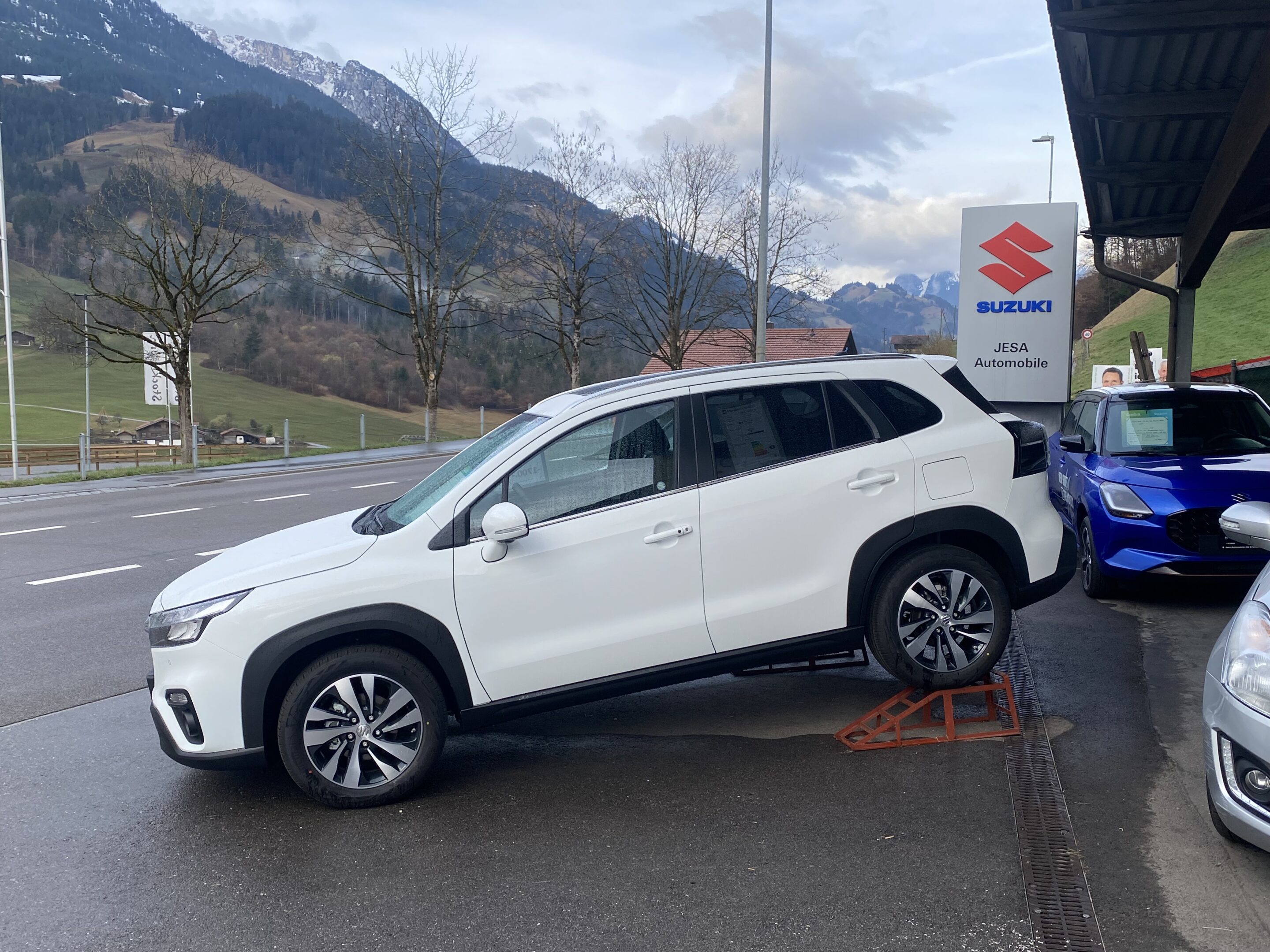 Weißer SUV vor Suzuki-Händler in bergiger Landschaft geparkt.