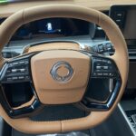 Luxuriöses Auto-Lenkrad in Lederoptik mit integrierten Bedienelementen und Display im Hintergrund.