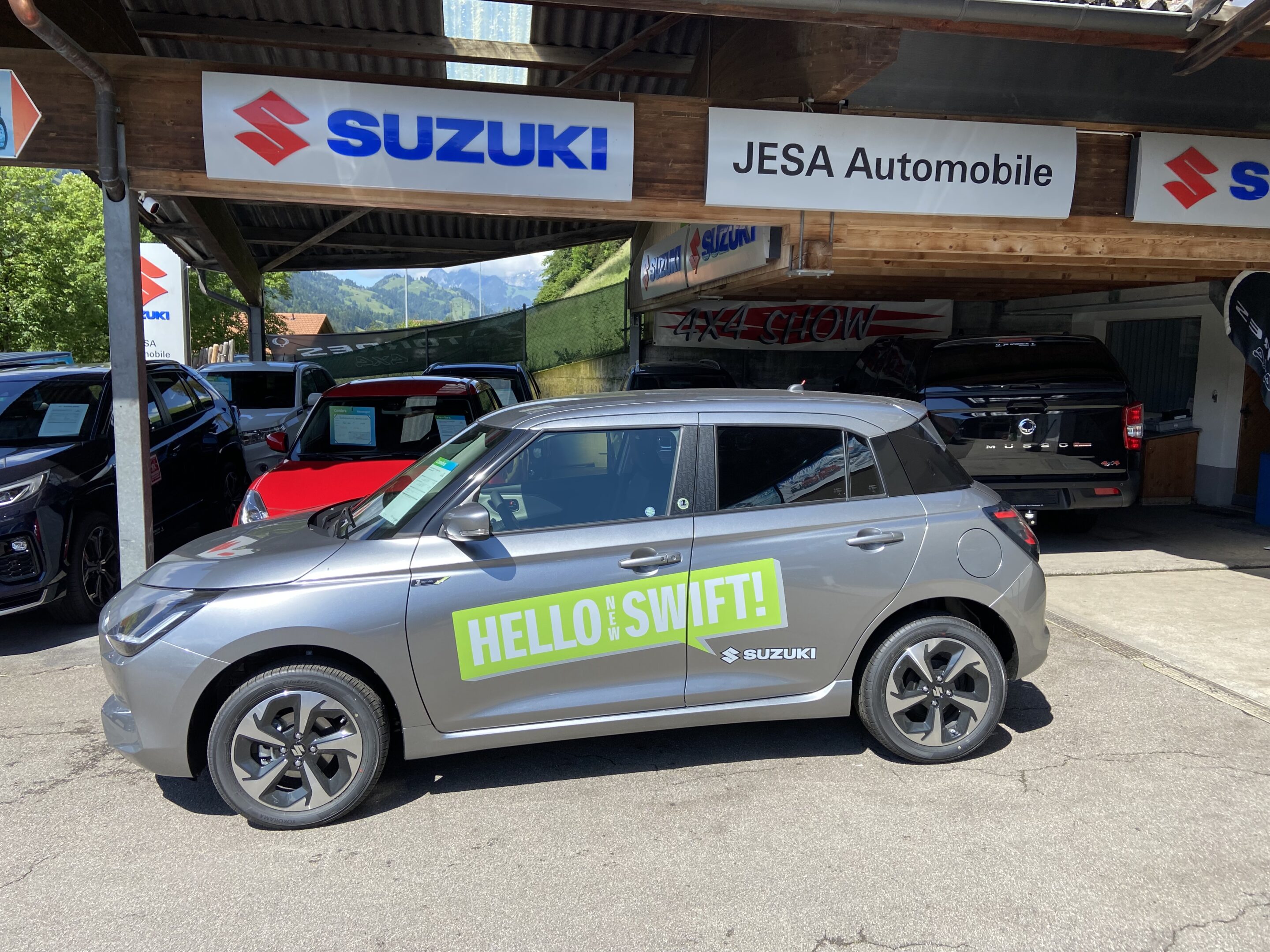 Silberner Suzuki Swift vor Jesa Automobile Händler mit Hello Swift Schriftzug, Parkbereich, Sonnenschein.