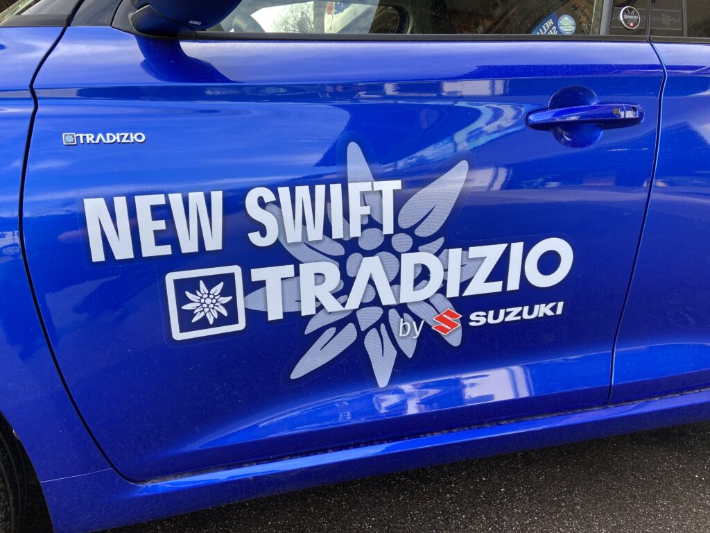 Blaues Auto mit Aufschrift New Swift Tradizio by Suzuki und Edelweiß-Logo auf der Seite.