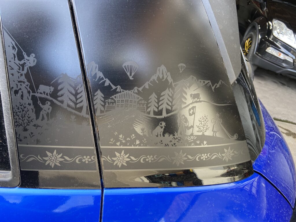 Autoaufkleber: Alpendekor mit Kletterern, Kühen, Chalet und Edelweiß auf getöntem Fenster eines blauen Fahrzeugs.