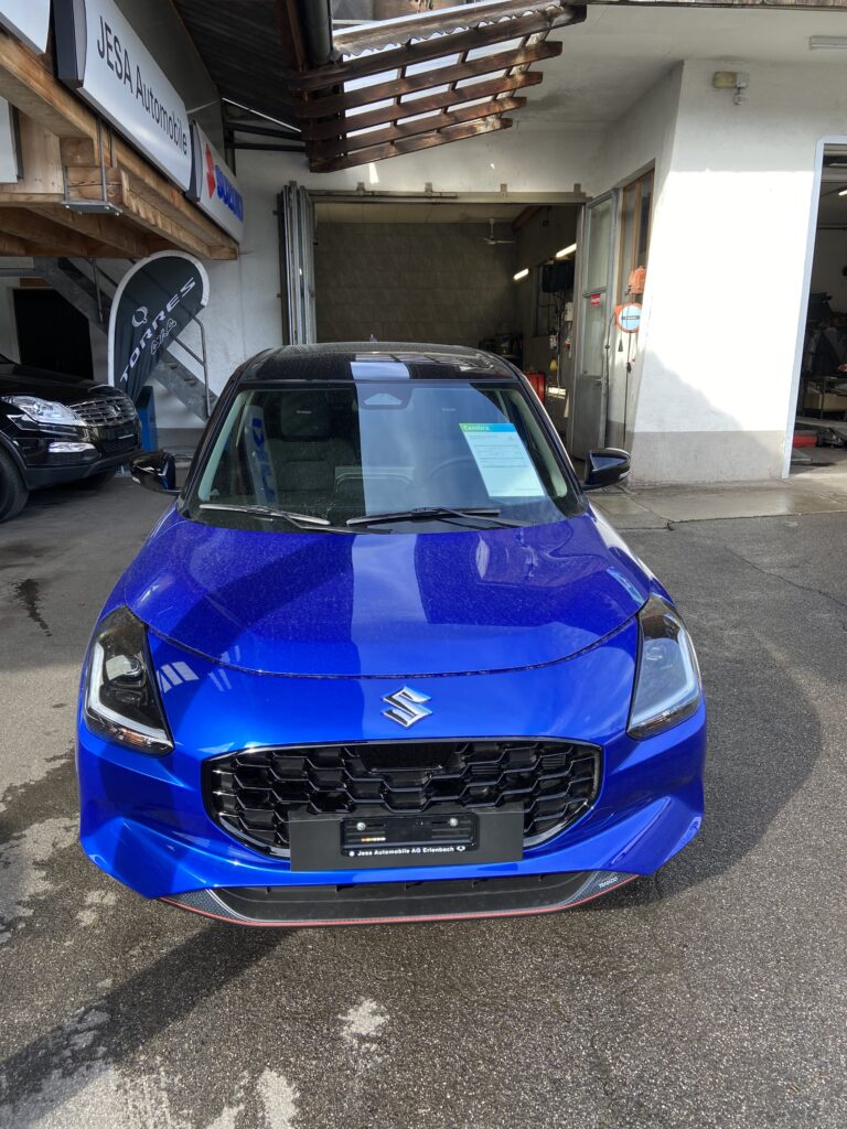 Blauer Suzuki vor einem Autohaus bei Jesa Automobile. Vorderansicht des Fahrzeugs, glänzend und modern.