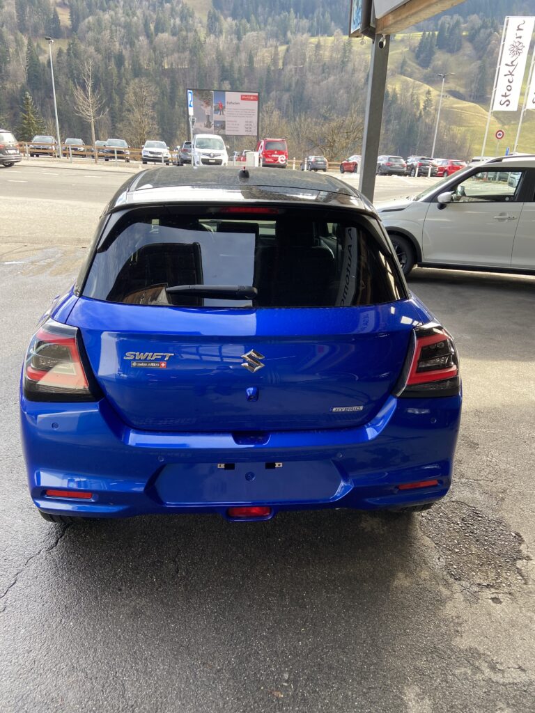 Blauer Suzuki Swift Hybrid geparkt auf nassem Parkplatz mit bergiger Landschaft im Hintergrund.