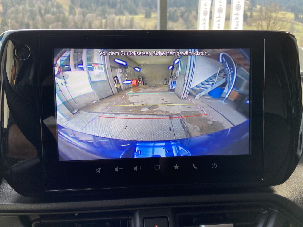 Rückfahrkamera-Ansicht des Autos zeigt eine Autowerkstatt mit Hebebühne und Leitlinien zum sicheren Zurücksetzen.