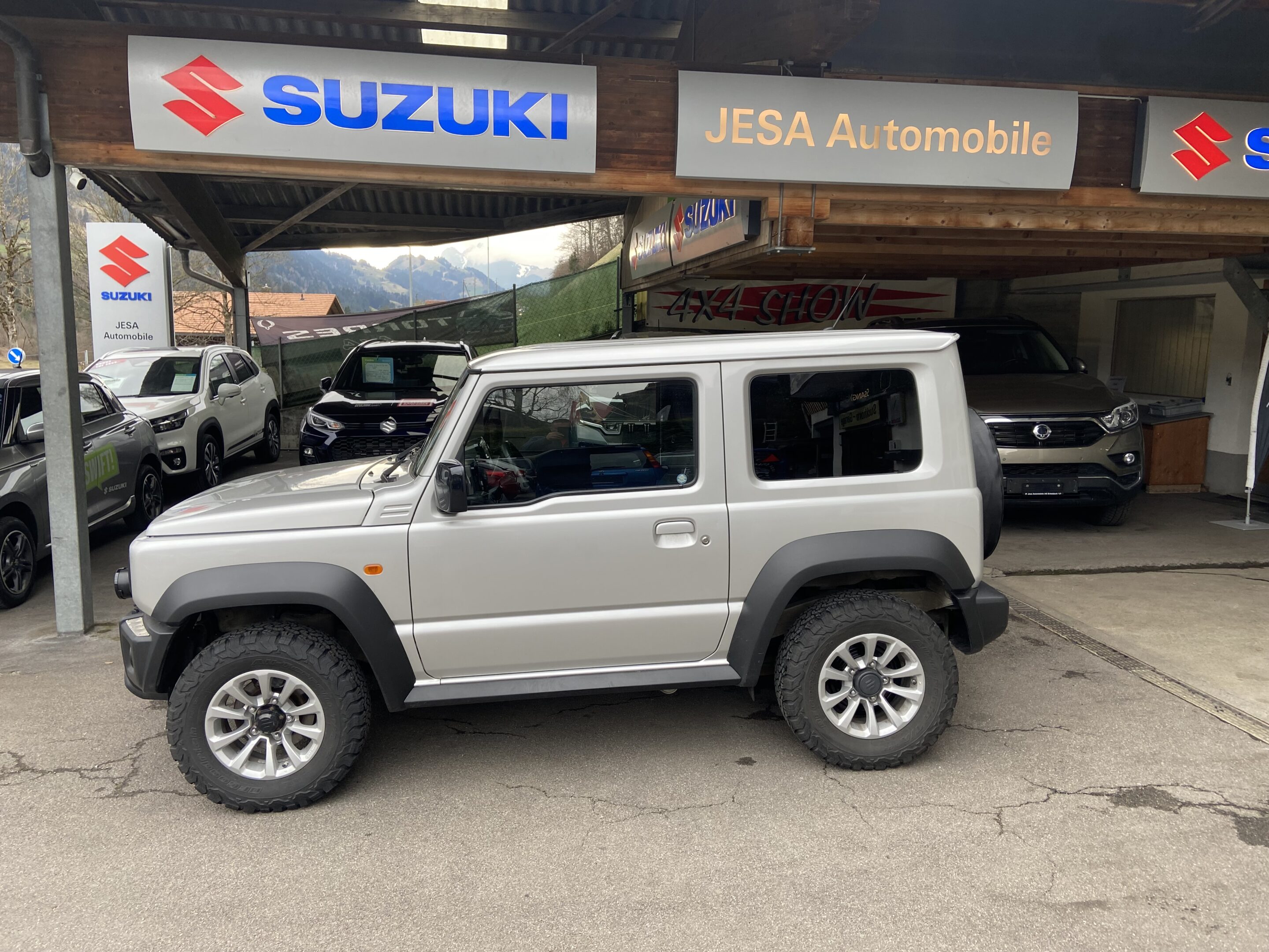 Silberner Suzuki Jimny geparkt vor einem Autohaus mit JESA Automobile Beschilderung.