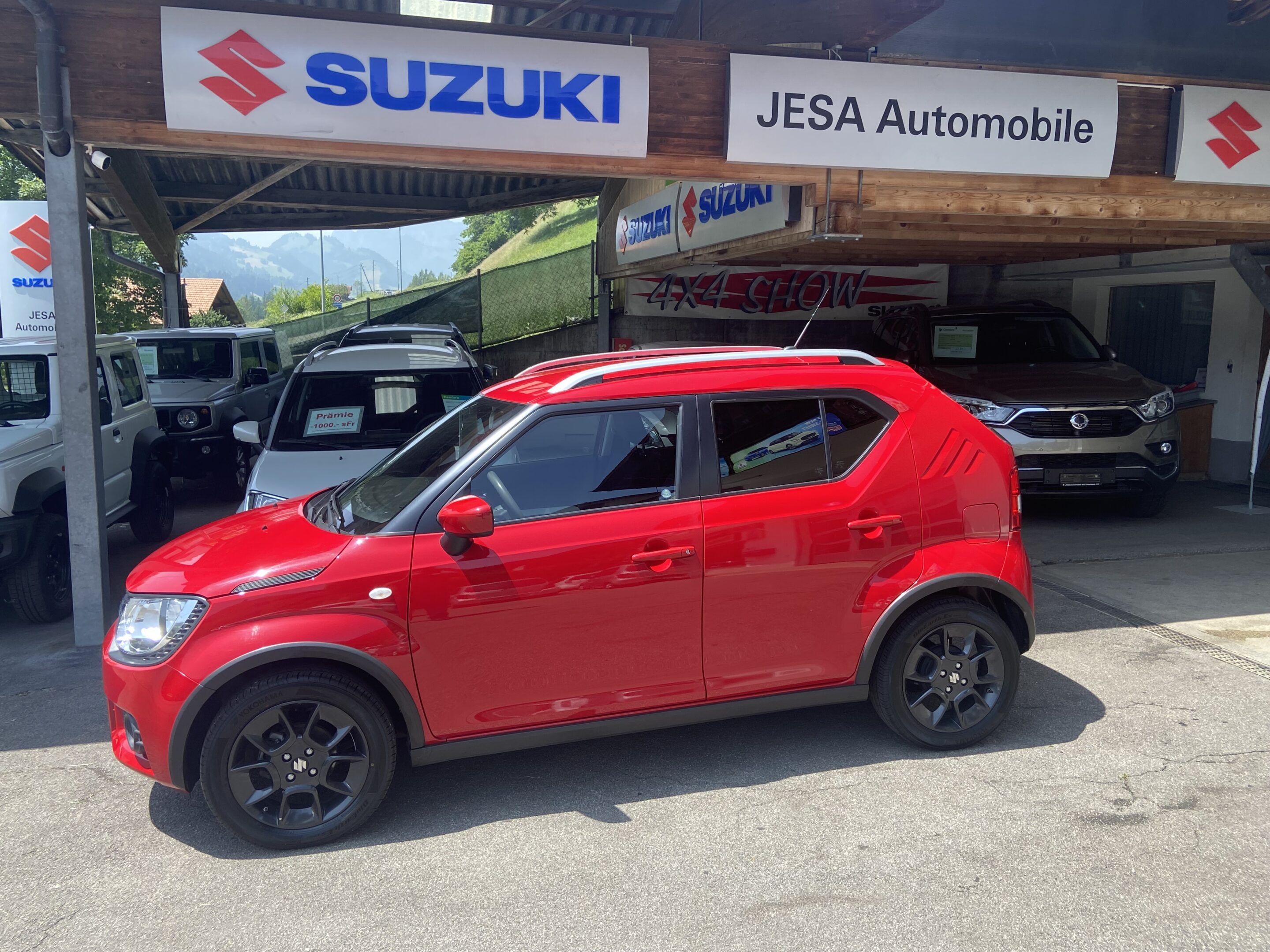 Roter Suzuki Ignis vor Jesa Automobile, umgeben von SUVs, bei sonnigem Wetter in einem Autohaus.