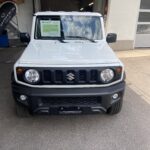 Weißer Suzuki Jimny vor einer Garage, Angebotsschild an der Frontscheibe sichtbar.