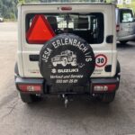 Heckansicht eines weißen Suzuki mit Werbebanner der JESA Erlenbach i/S Werkstatt, umgeben von grüner Landschaft.