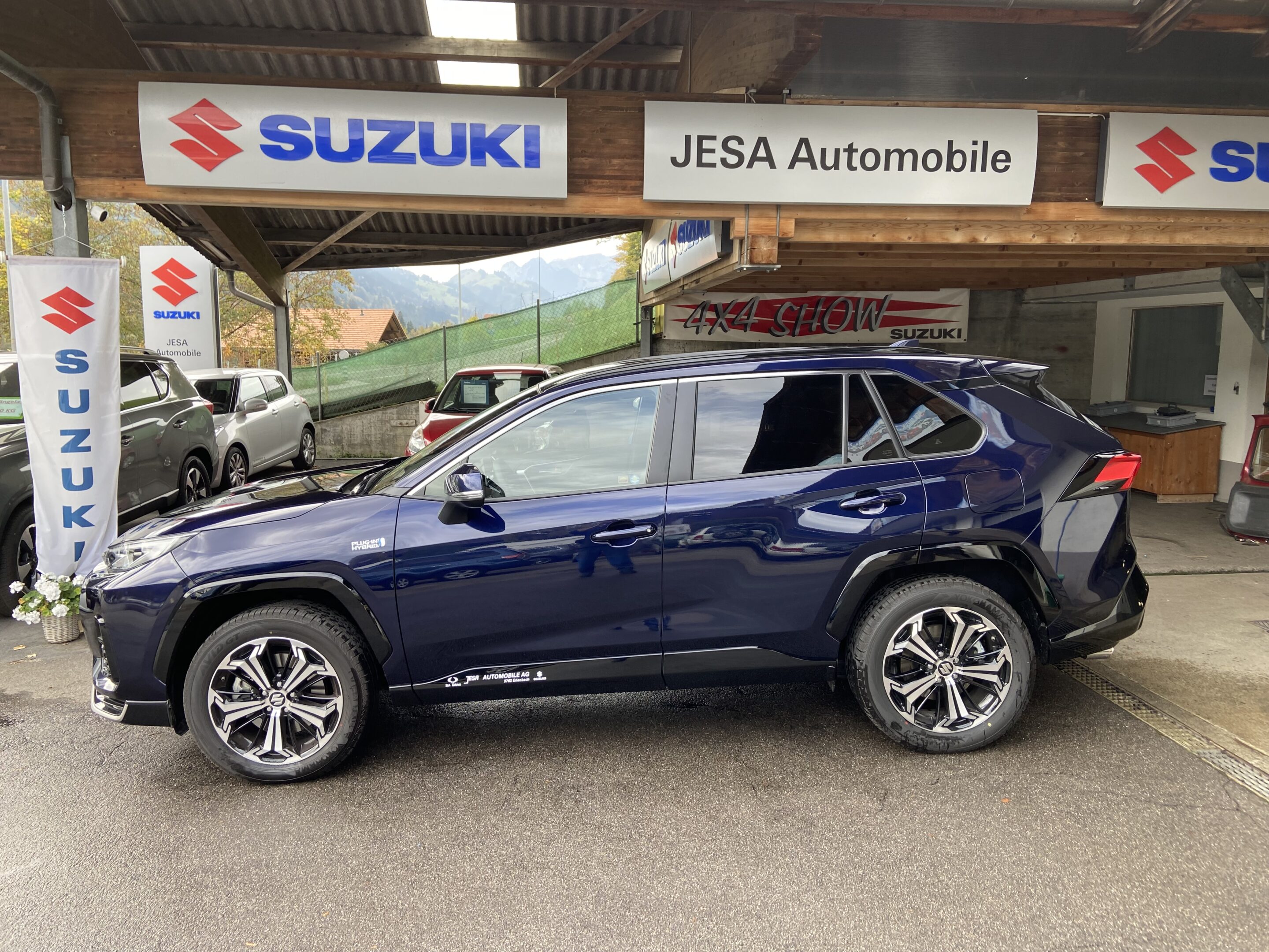 Blauer SUV vor einem Autohändler mit Suzuki-Schildern, im Hintergrund sind weitere Fahrzeuge zu sehen.