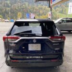 Rückansicht eines Suzuki Across Plug-in Hybrid vor Bergkulisse, geparkt bei einem Autohaus.