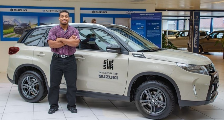 Mann steht lächelnd neben einem SUV im Autohaus mit Suzuki-Logo, bereit zur Probefahrt.