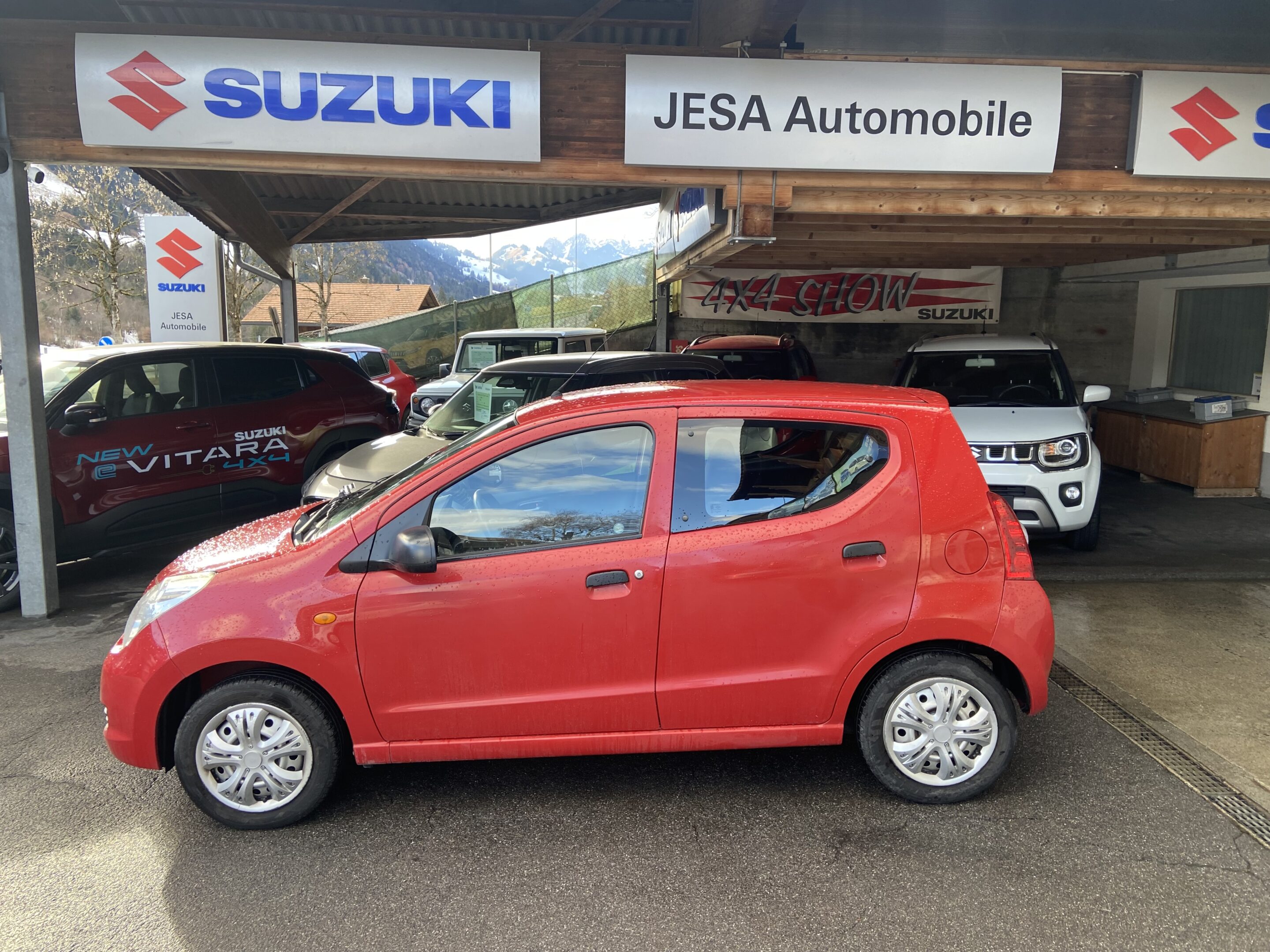 Rotes Auto vor Suzuki-Händler Jesa Automobile, umgeben von anderen Fahrzeugen, Berge im Hintergrund.