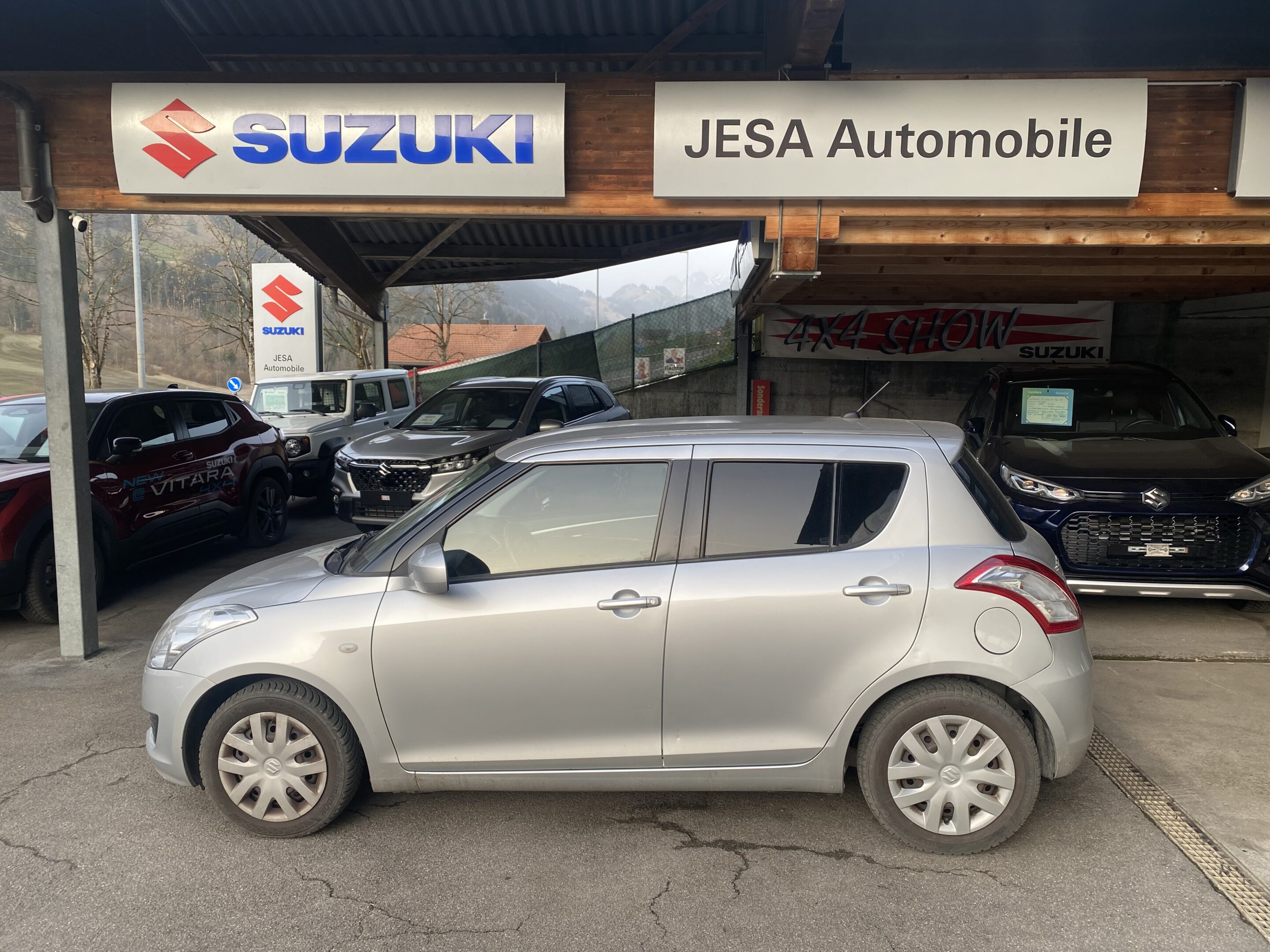 Silberner Suzuki Swift vor Jesa Automobil, umgeben von weiteren Suzuki-Modellen unter einem überdachten Carport.