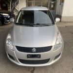 Silberner Suzuki Swift parkt vor einer Garage, Vorderansicht bei Tageslicht.