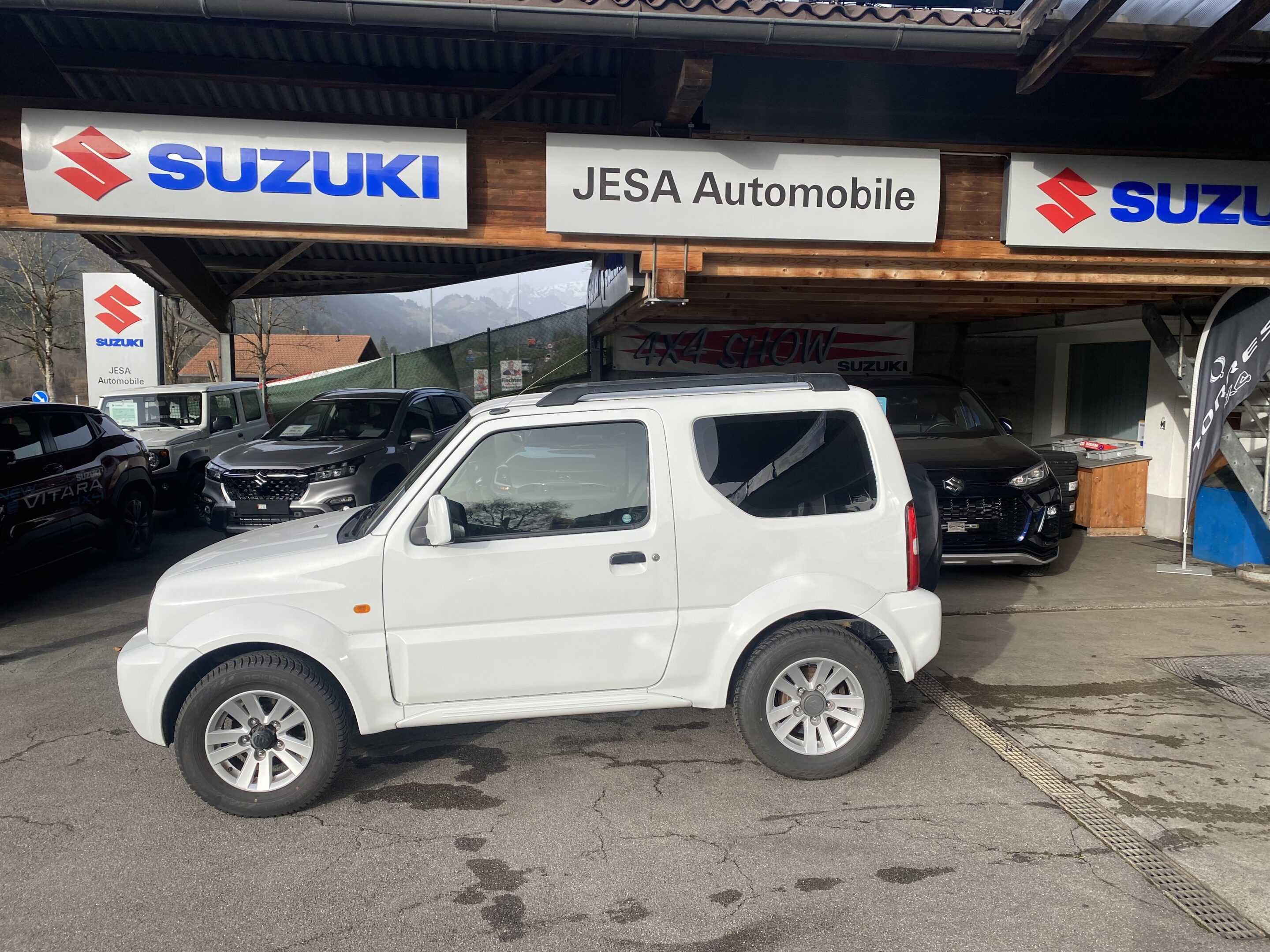 Weißer Suzuki Jimny vor JESA Automobile, umgeben von anderen Fahrzeugen, im Sonnenschein.