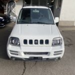 Weißer Suzuki Geländewagen vor Garage, frontal aufgenommen, bei Tageslicht. Ideal für Stadt- und Geländefahrten.