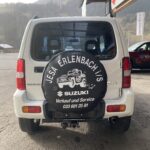 Suzuki SUV mit Ersatzradschutz von Jesa Erlenbach vor einem Autohändler. Verkauf und Service. Text und Logo sichtbar.