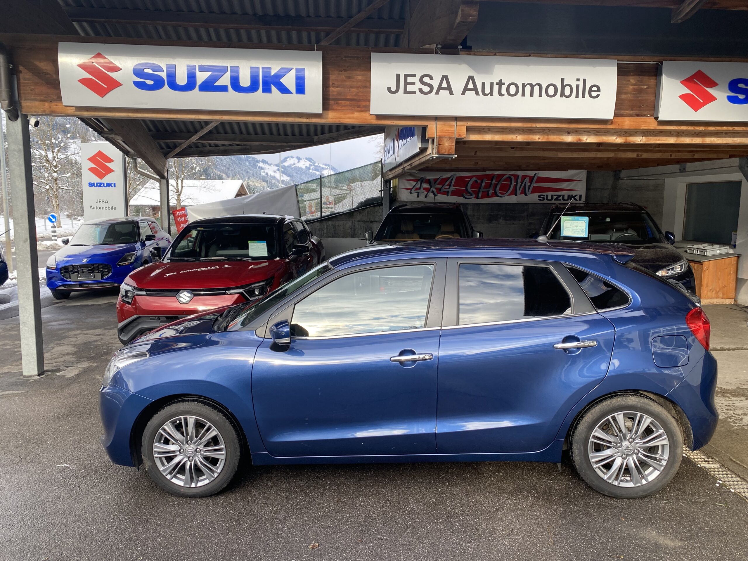 Blauer Suzuki vor Autohaus Jesa im Freien, mit mehreren anderen Suzuki-Modellen im Hintergrund, im Winter.