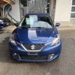 Blauer Suzuki Baleno vor einem Autohaus, Frontalansicht auf einem Parkplatz bei Tageslicht.