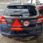 Blauer Suzuki Baleno mit 45er Schild und rotem Warnzeichen, geparkt auf Asphaltstraße im Freien.