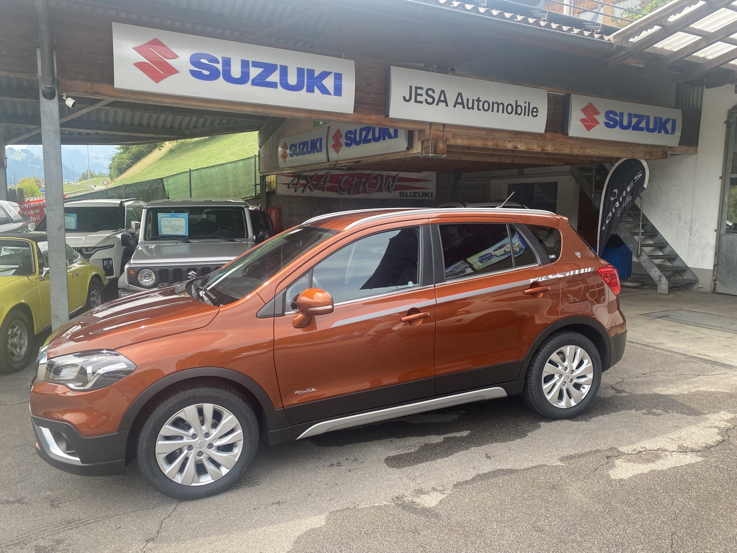 Orangefarbenes Suzuki SUV vor JESA Automobile Händler, umgeben von anderen Fahrzeugen, Suzuki-Logos sichtbar.