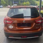 Roter Suzuki S-Cross mit CH-Aufkleber, geparkt auf einem Parkplatz mit umliegenden Fahrzeugen und bewaldeter Kulisse.