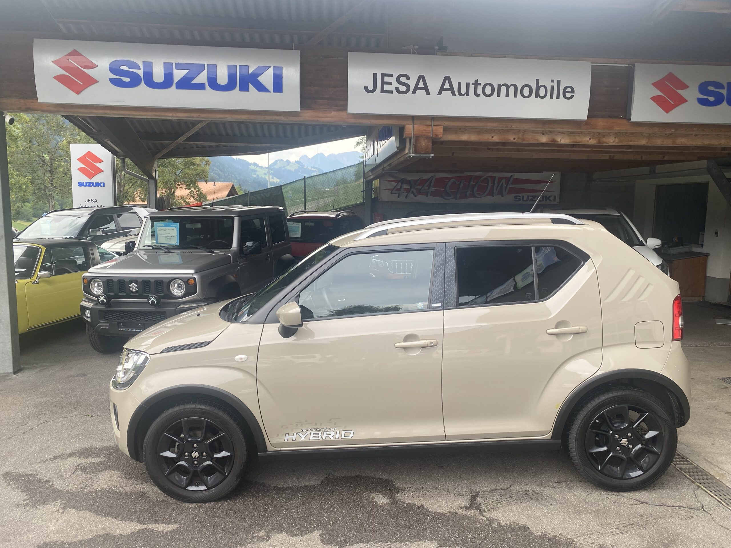 Beige Suzuki Hybrid Auto vor einem Autohaus mit Suzuki-Schildern im Hintergrund.