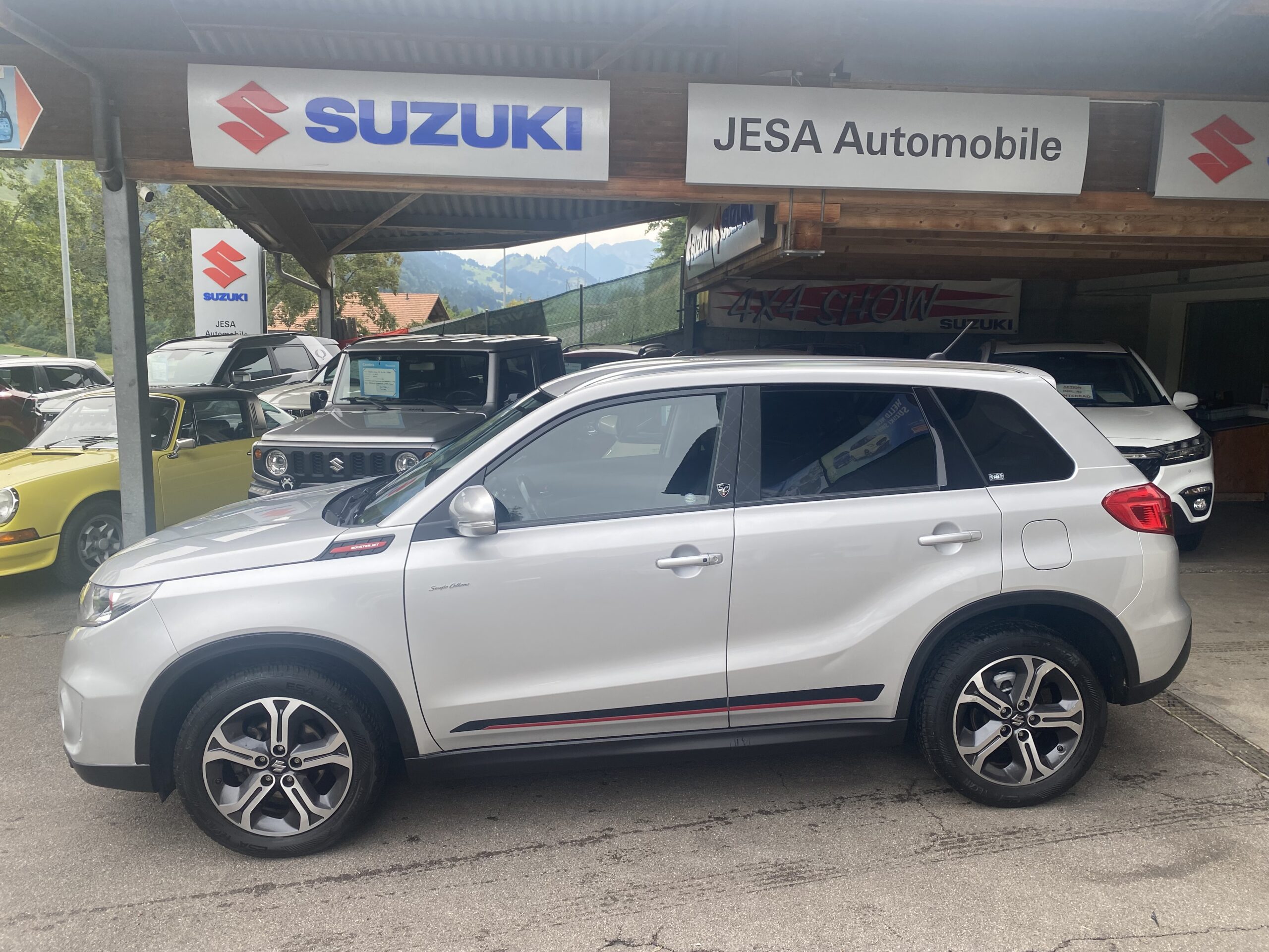 Silberner Suzuki SUV bei JESA Automobile, umgeben von Autos und Schildern, Ansicht von der Seite.