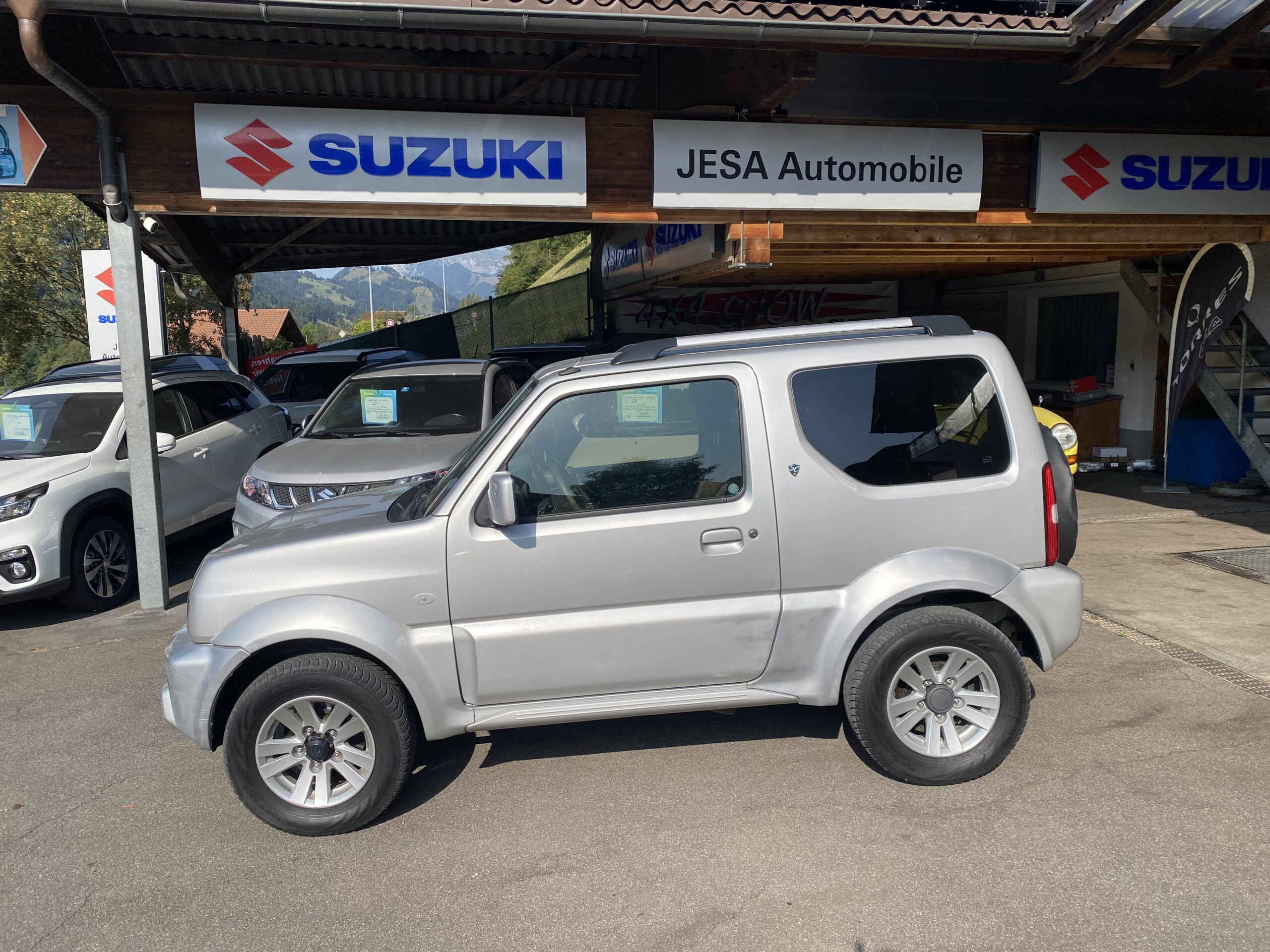 Silberner Suzuki Jimny parkt vor einem Autohaus mit Suzuki-Logo und Bergkulisse im Hintergrund.