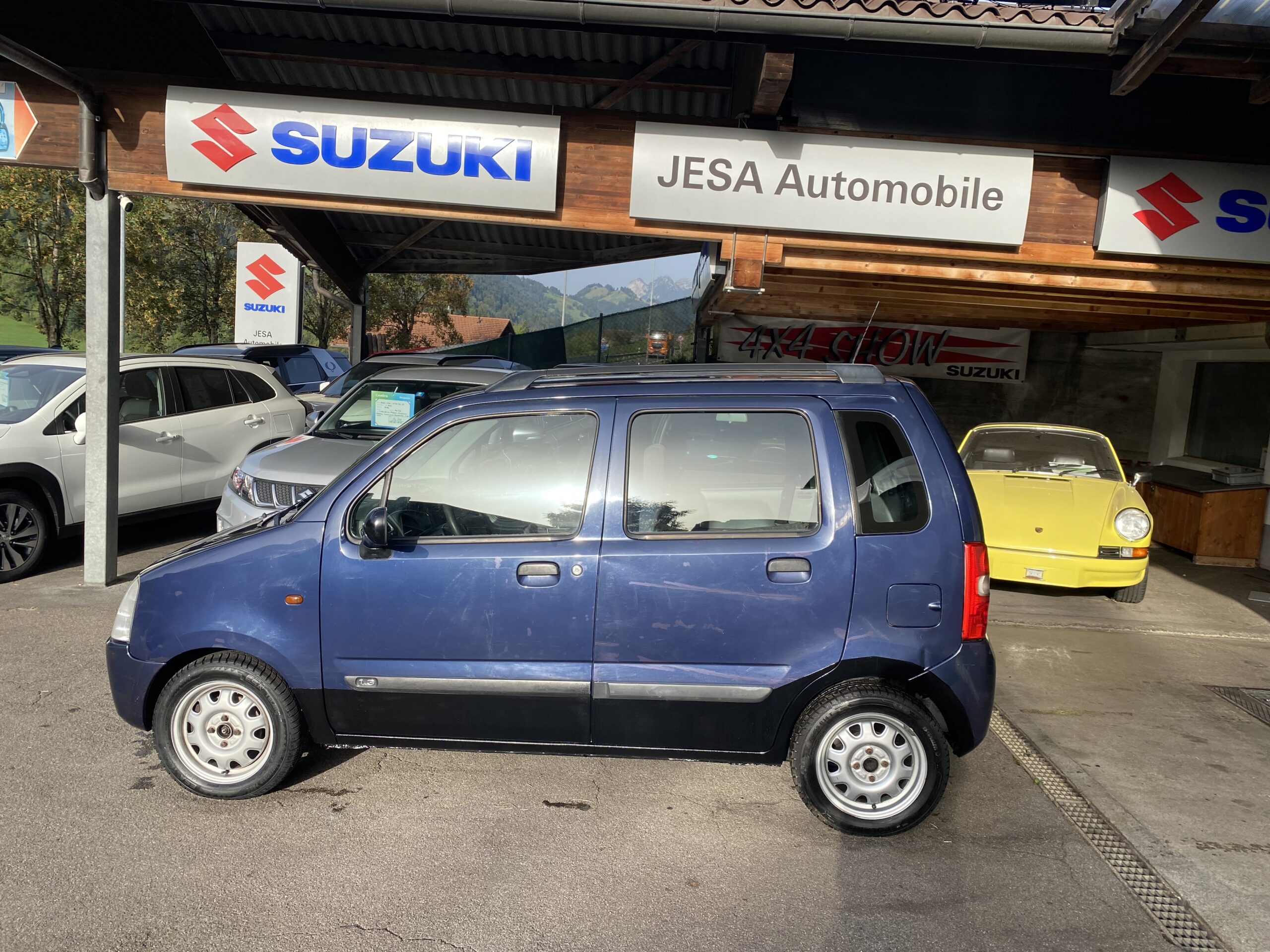 Blauer Suzuki Wagon R+ bei JESA Automobile vor Suzuki-Schild, mit einem gelben Oldtimer im Hintergrund.