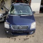 Blauer Suzuki Wagon R auf einem Parkplatz vor einer Werkstatt, im Hintergrund steht ein rotes Auto.