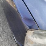 Kratzer und Delle an blauem Autostoßfänger neben Scheinwerfer auf Asphalt.