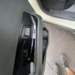 Autotür mit Bedienknöpfen für Fensterheber; geöffnete Tür zeigt Schuhwerk auf Asphalt.