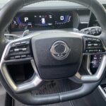 Lenkrad eines modernen Autos mit eingebauter Steuerung und digitalem Armaturenbrett im Hintergrund.