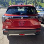 Roter Suzuki S-Cross Hybrid auf Parkplatz, umgeben von grüner Landschaft und blauen Himmel im Hintergrund.