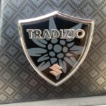 Schwarzes Tradizio-Emblem mit Edelweißblume, silbernem Rahmen und Hintergrundmuster.
